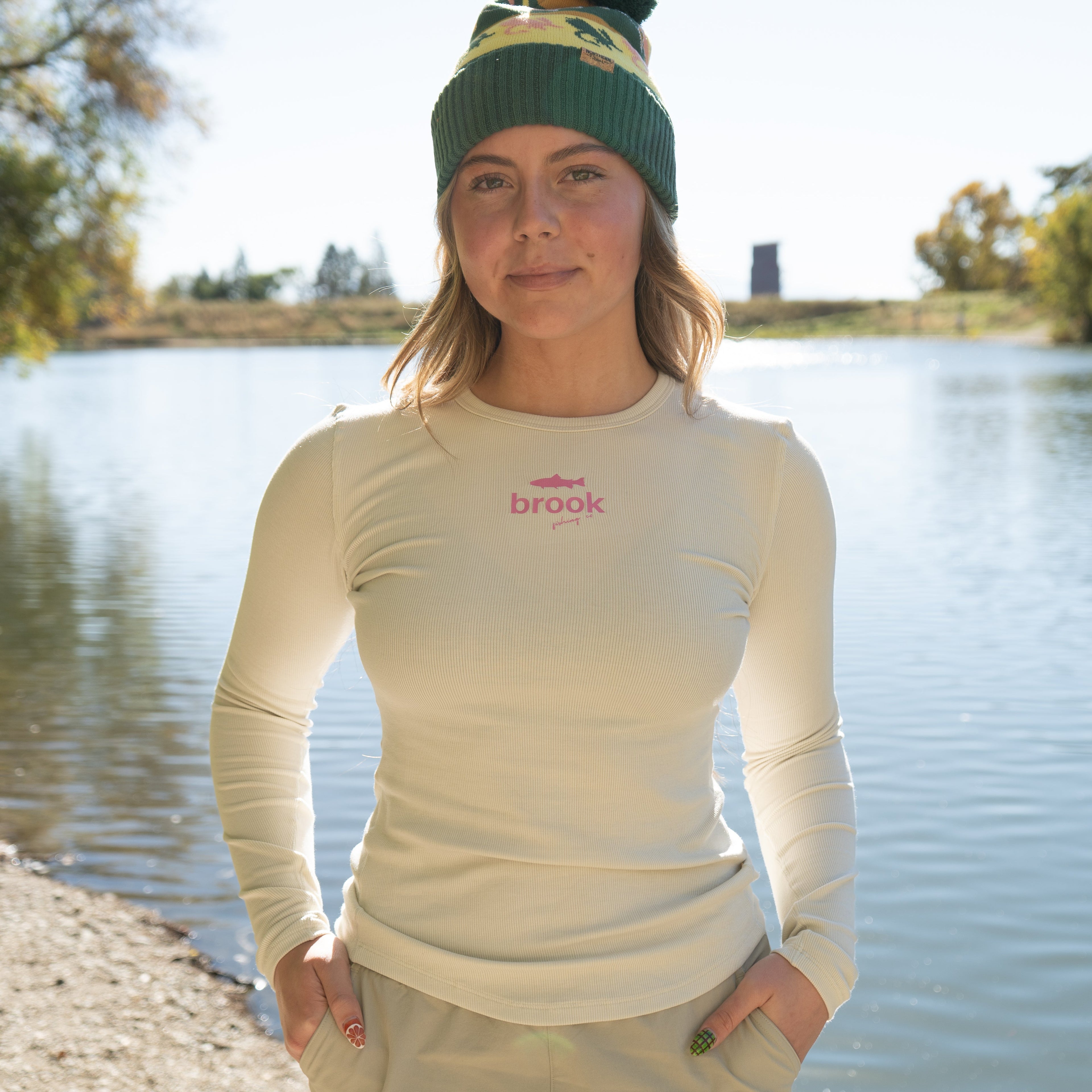 Woman wearing cream ribbed modal fishing tee with pink logo, standing near river.