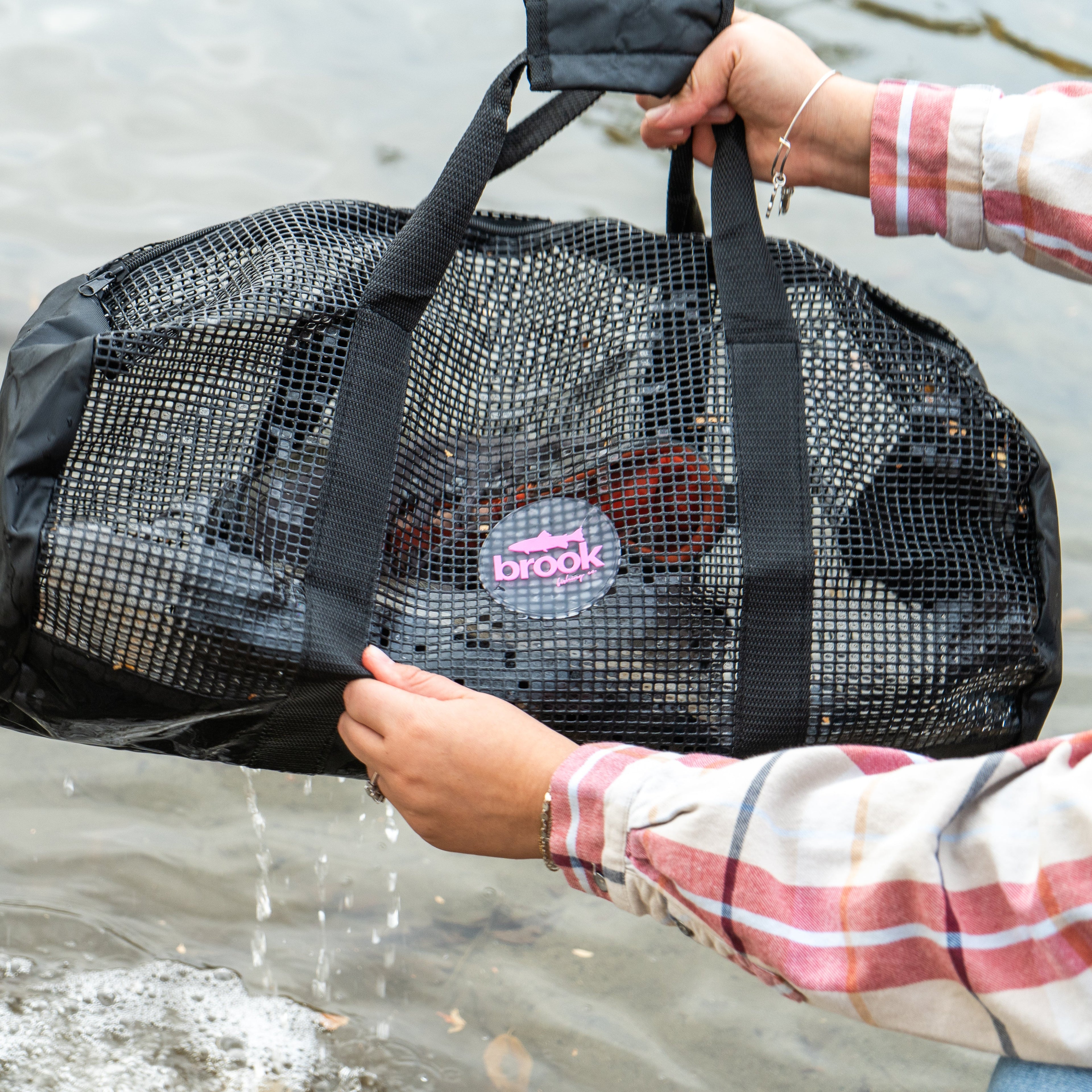 Person rinsing wader boots in a black mesh gear bag in shallow water, with the pink Brook Fishing Co. logo visible and water flowing through the mesh.