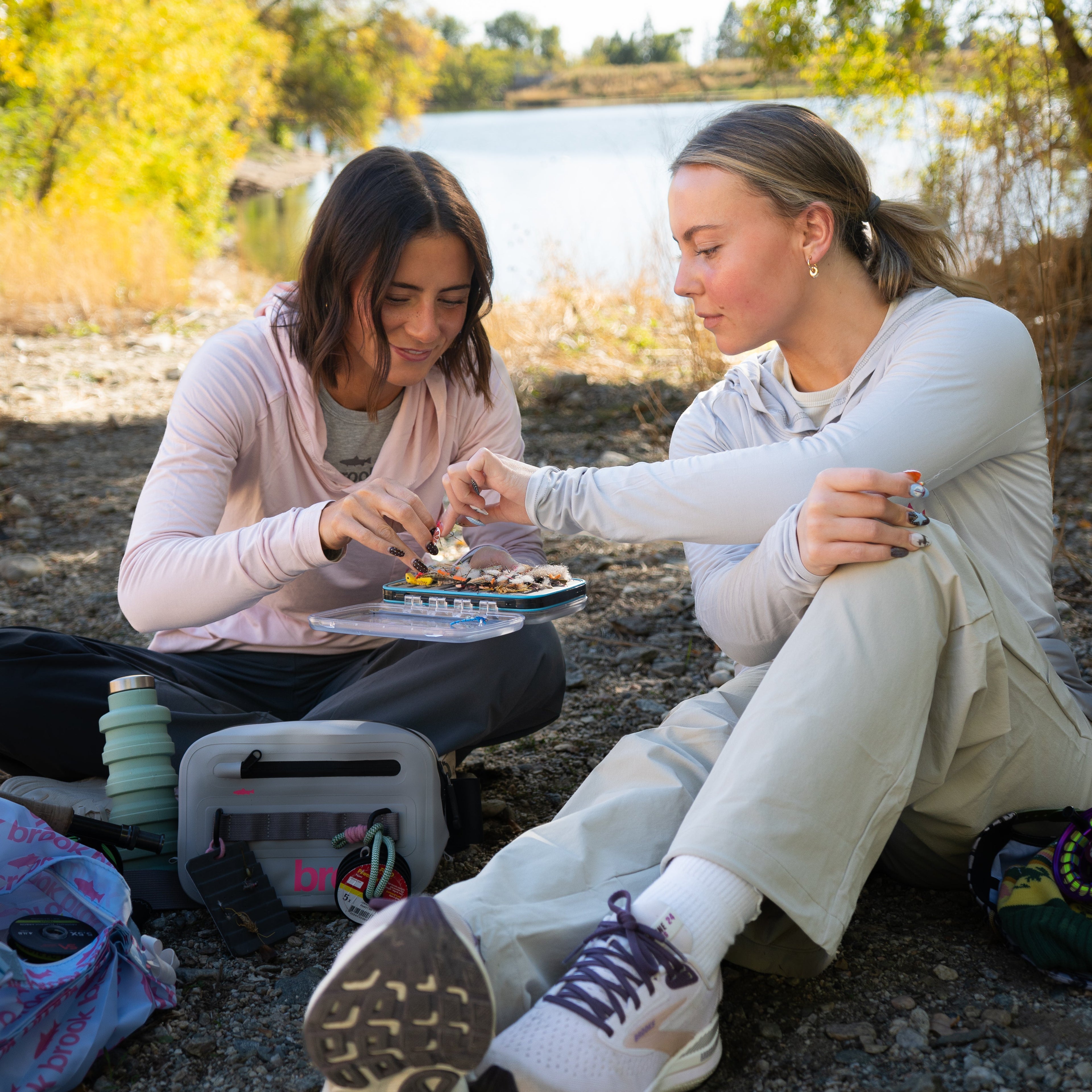 Two female anglers sitting near the river, passing a fly while wearing Pink Brook Bamboo Sun Hoodies with ponytail openings.