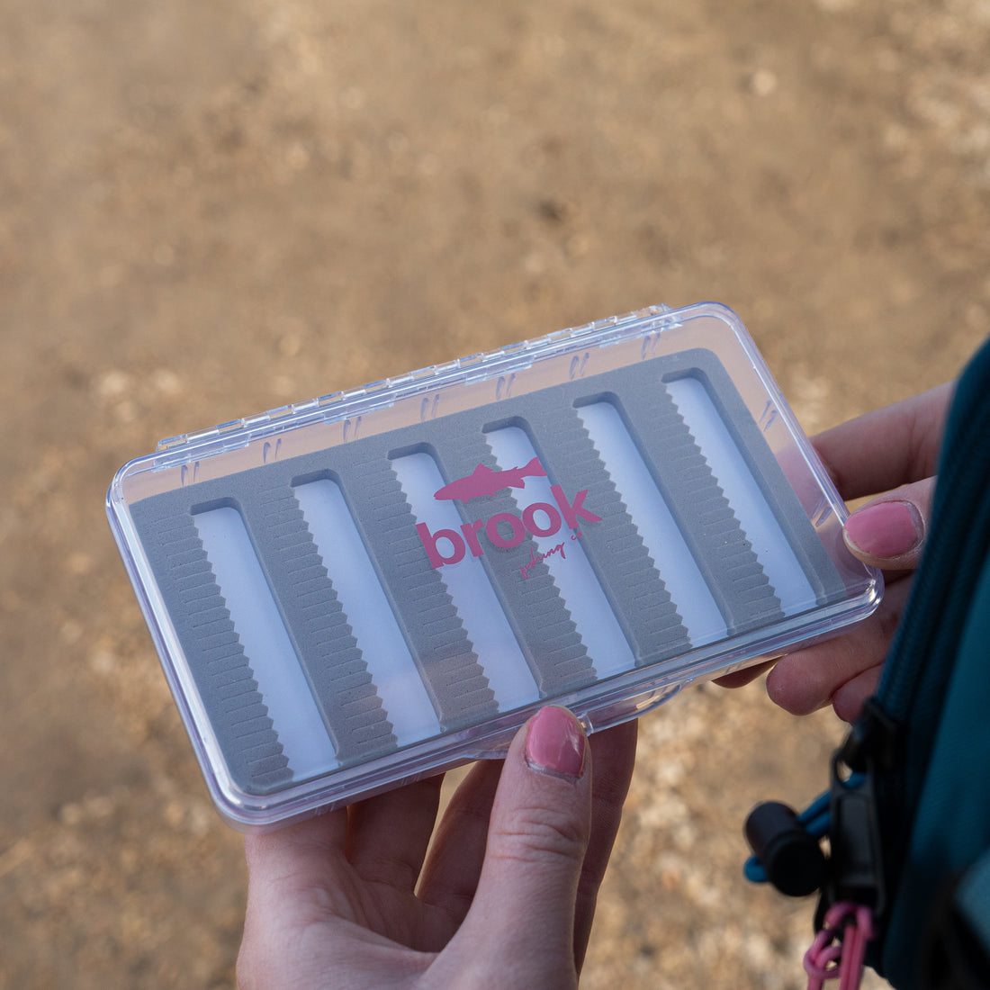 Hands holding a Brook Fishing Co. slim fly fishing box with clear lid and gray foam slots designed to organize trout flies on the river.