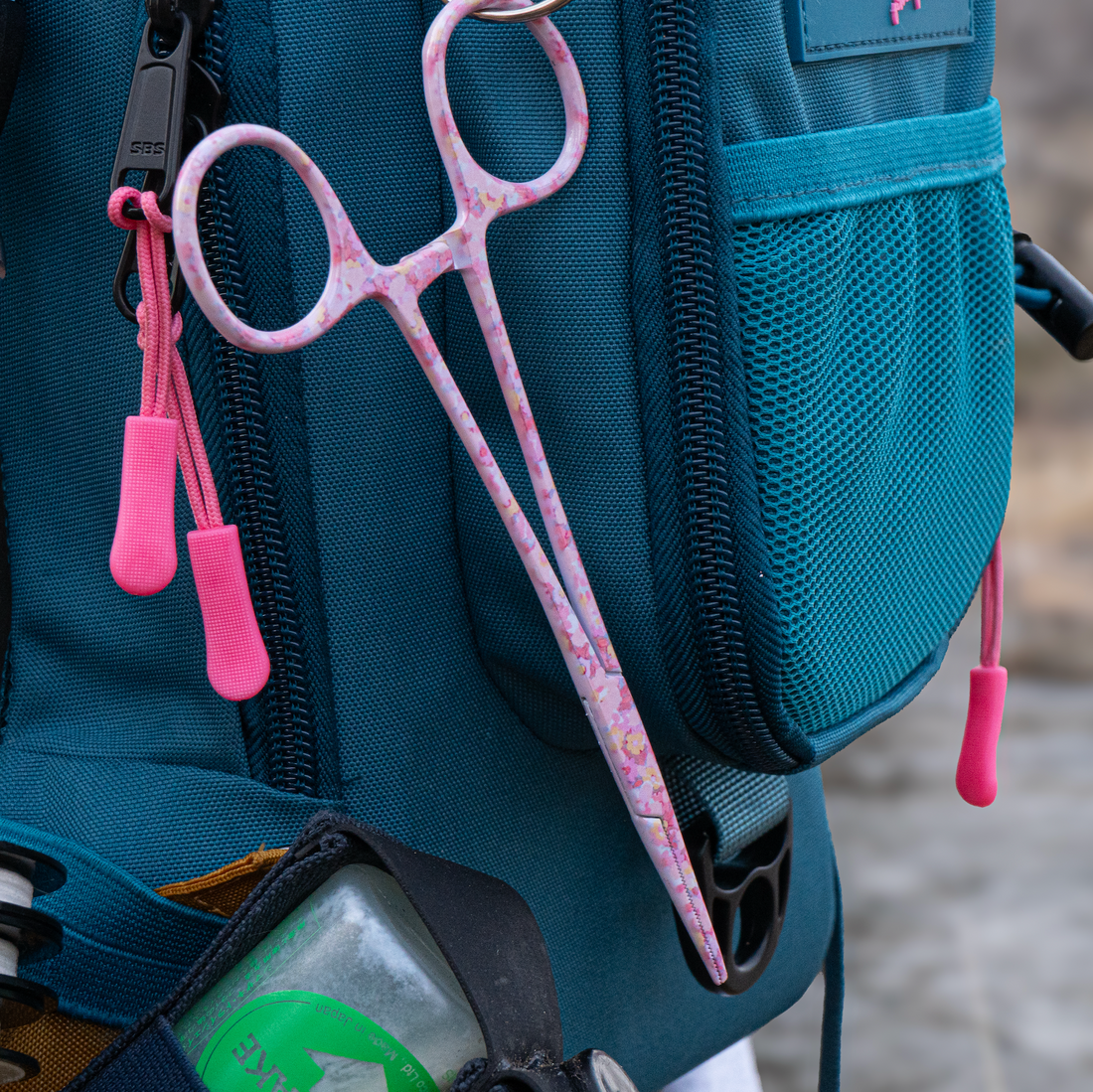 Pink fly fishing scissors attached to a teal backpack with a blurred natural background