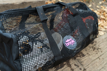 “Black mesh gear bag with the Brook Fishing Co. pink logo, shown outdoors on wooden steps with fallen leaves. The bag is filled with wet fishing boots and gear visible through the mesh.”