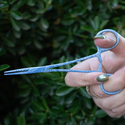 Angled view of hand holding Mini Waves fishing hemostats, highlighting stainless steel finish and soft wave pattern.