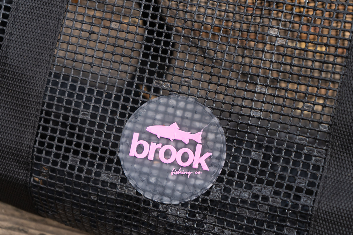 “Close-up of the Brook Fishing Co. pink logo on the black mesh gear bag, showing the trout icon and branding against the open-grid PVC mesh.”