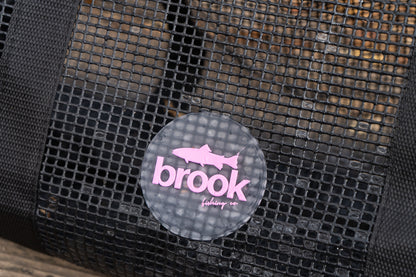 “Close-up of the Brook Fishing Co. pink logo on the black mesh gear bag, showing the trout icon and branding against the open-grid PVC mesh.”