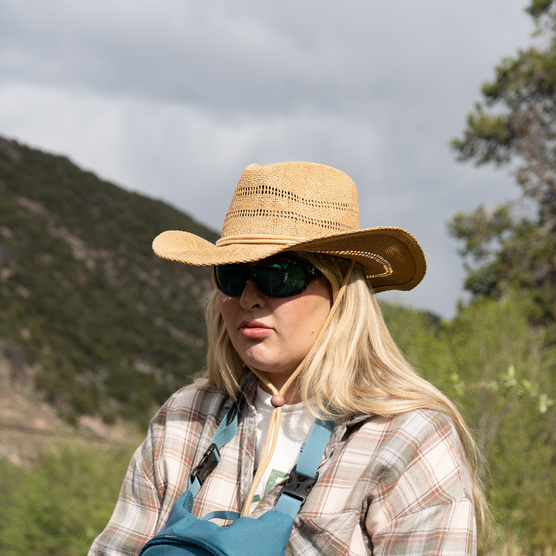 Person wearing a straw hat and sunglasses in a natural setting with mountains and trees.